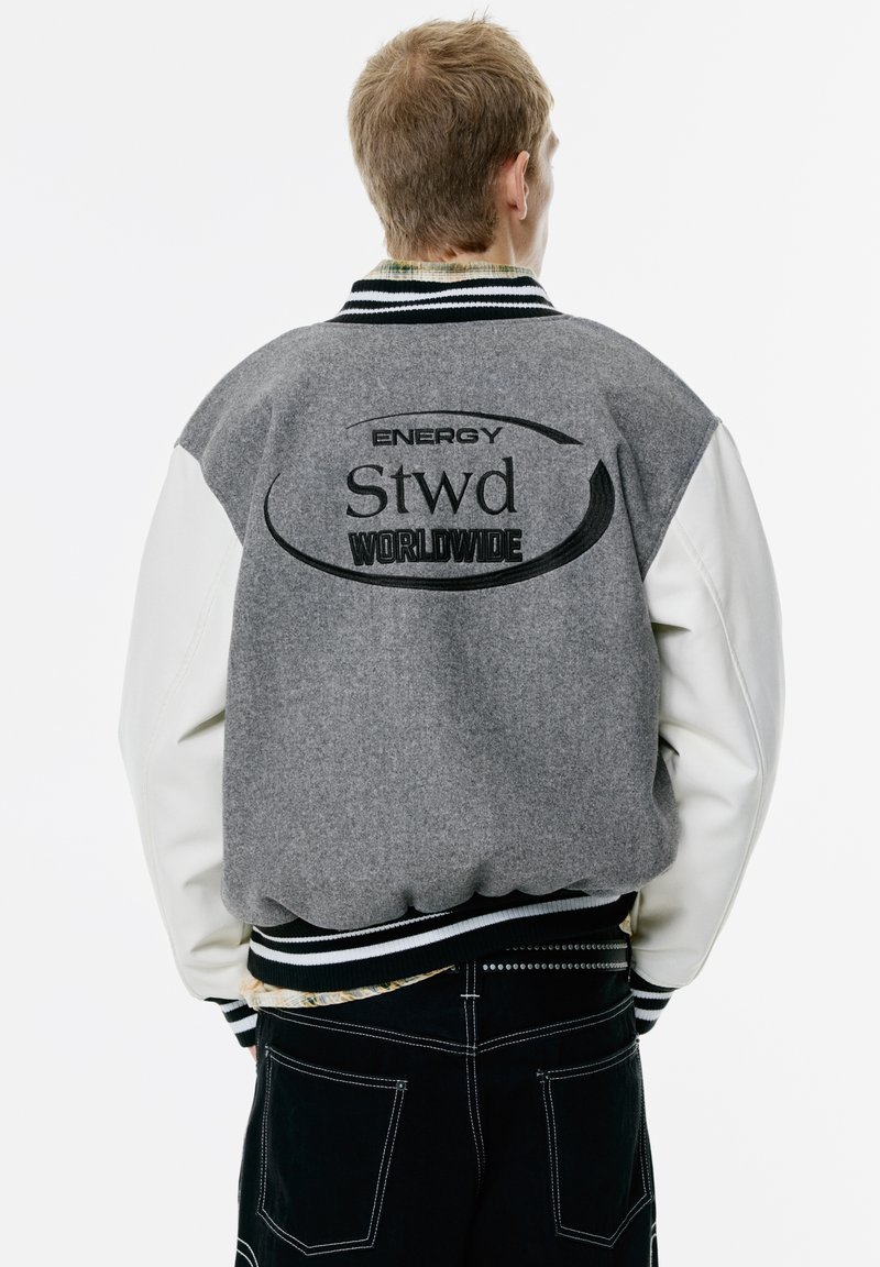 Bomber en mélange de laine gris avec des manches en cuir blanc, col et poignets côtelés. Présente un texte logo noir brodé dans le dos.