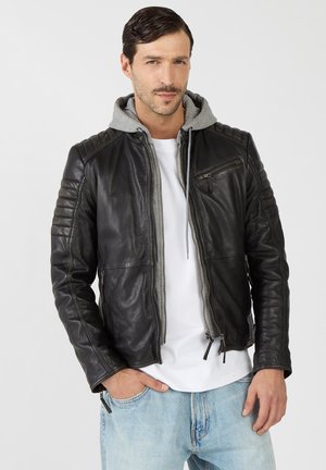 Man draagt een zwarte leren jas met doorgestikte mouwen over een grijze hoodie en wit overhemd, staand met één hand in de zak van lichtblauwe spijkerbroek.