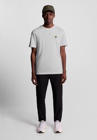 Lichtgrijs T-shirt met een klein geel logo op de borst, gecombineerd met zwarte broek en een groene pet. Sneakers zijn licht van kleur met witte accenten.
