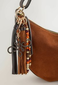 Coin de sac en daim marron avec fermeture éclair dorée, bandoulière en cuir noir et pompons attachés ainsi que porte-clés avec perles colorées.