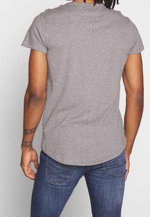 Personne portant un t-shirt gris ajusté à manches courtes et un jean bleu, vue de dos, avec des tatouages visibles sur les bras.