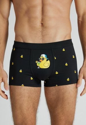 Mandlig torso iført sorte boxershorts med mønster af gule gummiduer, en stor due holder en cocktail, iført solbriller og kasket.