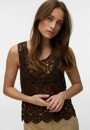 Kvinde med langt brunt hår iført en ærmeløs mørkebrun hækletrøje og lyse beige bukser, stående mod en ensfarvet baggrund.