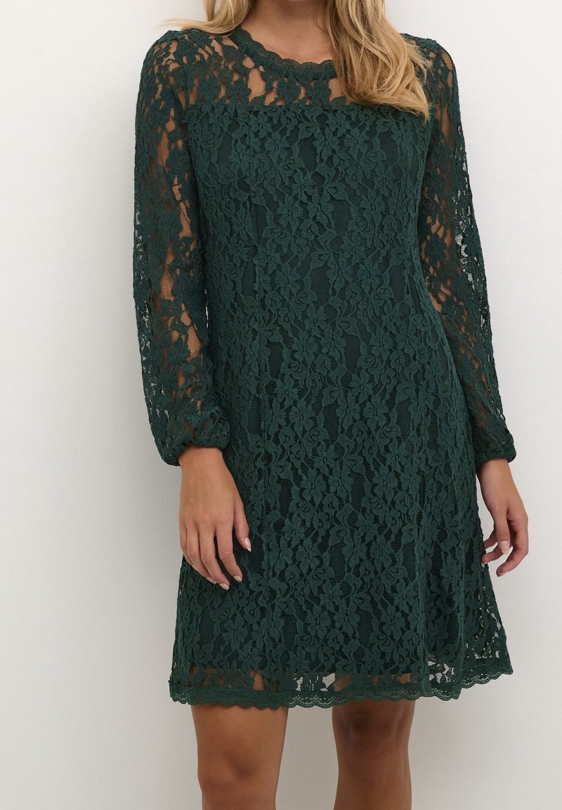 Femme portant une robe longue à manches longues en dentelle florale vert foncé avec un ourlet festonné, debout devant un fond clair uni.