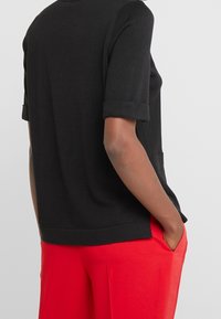 Czarny t-shirt z krótkim rękawem i podwiniętymi mankietami, w luźnym fasonie, zestawiony z czerwonymi spodniami. Posiada boczną kieszeń dla zwiększenia funkcjonalności.