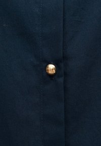 Πουκάμισο με κουμπιά από ύφασμα ναυτικού μπλε, με λεπτομέρεια από λαμπερό χρυσό κουμπί, ομαλή υφή και τακτοποιημένα ραφικά κατά μήκος της φάσας κουμπιών.