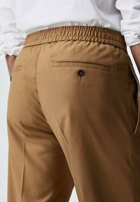 Braune Hose mit einem elastischen Bund, ausgestattet mit zwei Gesäßtaschen mit Knopfverschluss und einer glatten Textur.