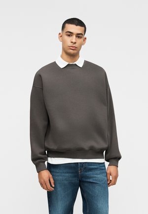 Mann trägt dunkelgrauen, übergroßen Pullover über weißem Hemd mit Kragen und blauen Jeans, steht vor einfarbigem hellem Hintergrund.