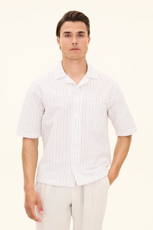 Jonge man met donker haar, gekleed in een wit overhemd met korte mouwen met dunne verticale strepen en lichte broek tegen een effen achtergrond.