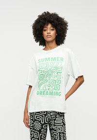 T-shirt oversize blanche avec impression graphique verte portant les mots "RÊVER D'ÉTÉ" et comportant des éléments de design tropicaux. Pantalon noir à motifs abstraits.
