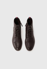 Botas de tobillo de cuero marrón con punta redondeada, cordones y una cremallera lateral. Presenta un acento de tela a rayas en el talón. Textura suave.