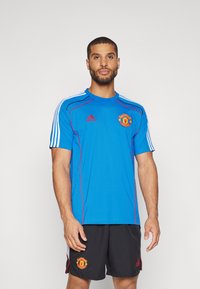 Mėlynas sportinis marškinėlis su trumpomis rankovėmis, raudonais akcentais, Adidas logotipu ir Mančesterio „United“ emblema ant krūtinės, derinamas su juodais šortais.