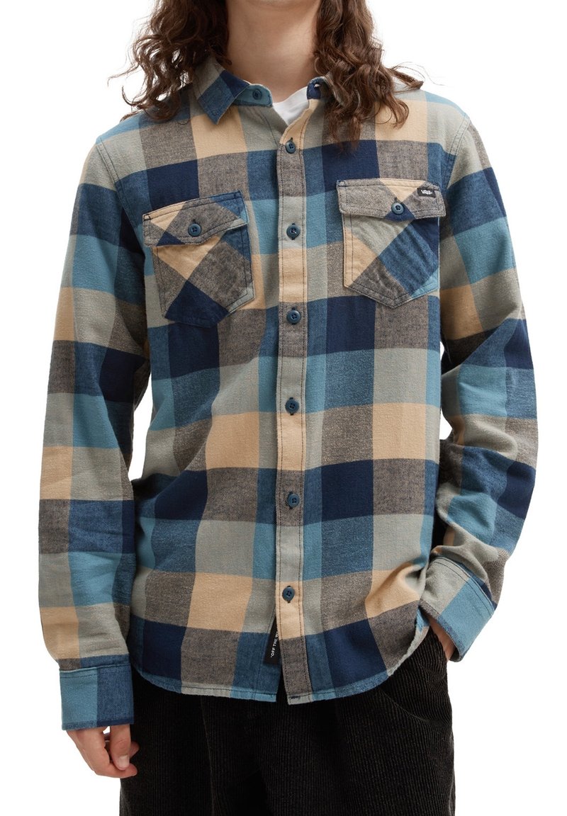 Personne portant une chemise à carreaux bleu, beige et gris à boutons, avec deux poches poitrine avant, une main dans la poche, sur fond blanc.