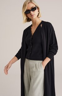 Długi czarny kardigan na ciemnym prążkowanym topie z dekoltem w serek, zestawiony z jasnoszarymi plisowanymi spodniami. Gładka faktura, luźny krój, bez widocznych zapięć.