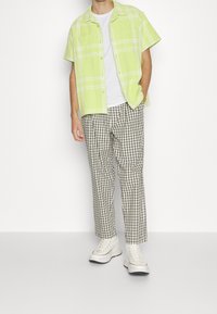 Man wearing light green short-sleeve plaid shirt over white T-shirt, beige checkered pants, and white platform sneakers standing against white background.