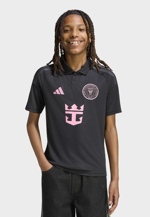 Šypsosi jaunas žmogus su kasomis, dėvintis juodą polo marškinėlį su rožiniu Adidas logotipu ir Club Internacional de Futbol Miami emblemą.