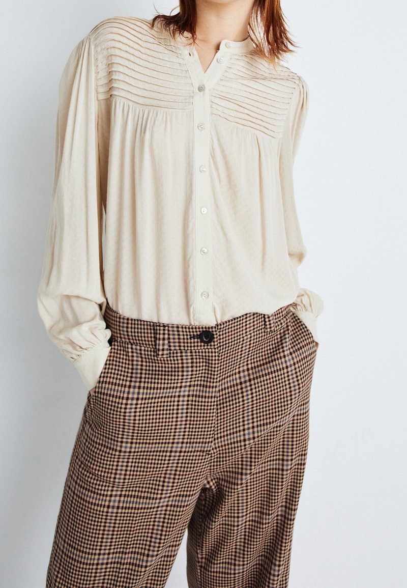 Donna che indossa una camicia a pieghe color crema con bottoni, infilata in pantaloni a vita alta a quadri marroni e neri, con le mani nelle tasche.