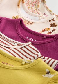 Cuatro camisetas de bebé acanaladas en crema floral, morado, crema con rayas marrones y mostaza con pequeños lazos, colocadas en capas sobre un fondo blanco.