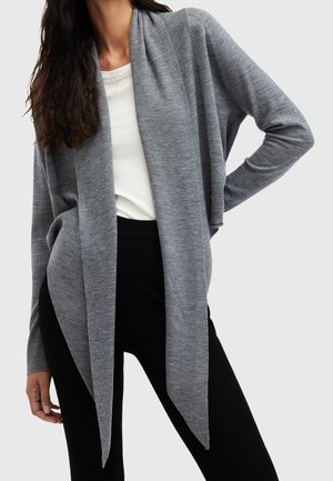 Kvinde iført en lang grå cardigan med åben front over en hvid skjorte og sorte bukser, stående med den ene hånd på hoften.