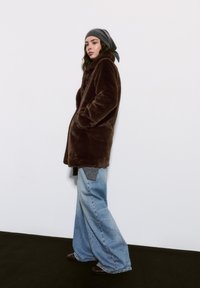 Manteau en fausse fourrure marron avec poches latérales, coupe oversize, manches longues, superposé sur un pull gris et associé à un jean en denim à jambes larges.