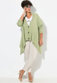 Ljusgrön cardigan med V-ringning, svarta knappar och en flytande design. Kombinerad med en vit skjorta och beiga byxor, avslutad med nakna klackar.