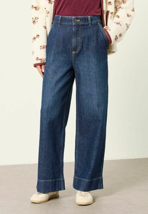 Frau trägt dunkelblaue Jeans mit weitem Bein, beige Sneakers, ein rotes Top und eine weiße Strickjacke mit kleinen Blumenmustern.