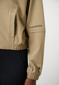 Beige jakke med elastisk mansjett og svart "CHAMPION" tekst over albuen, brukt over svarte bukser.