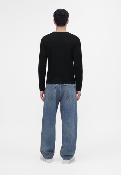 Homme portant un haut à manches longues noir, un jean bleu ample et des chaussures blanches, debout de dos sur un fond blanc uni.