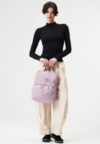 Hellblauer Rucksack aus strukturiertem Stoff, mit zwei oberen Tragegriffen und subtilen Details. Getragen von einem Modell in schwarzem Oberteil und cremefarbener Hose.