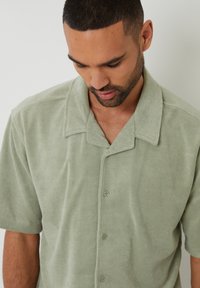 Chemise vert clair à manches courtes en tissu doux et texturé avec un col, dotée d'un bouton sur le devant et d'une coupe décontractée.