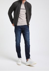Giacca zip nera su un pullover beige texturizzato a righe, abbinata a jeans blu scuro e sneaker bianche.