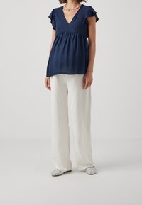 Navy blue V-neck top with ruffled sleeves, paired with white wide-leg pants and sparkly slip-on shoes. Smooth fabric with draping.