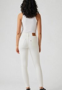 Jeans blancos ajustados con una tela texturizada, corte a media altura en la cintura y bolsillos traseros. Cuenta con un parche de marca de cuero marrón en la cinturilla.