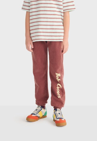 Niño con camiseta de manga corta a rayas, pantalones deportivos marrones con el texto "Bob Closes" y zapatillas de colores, de pie contra un fondo liso.