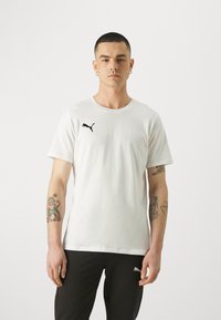 Baltos medvilnės marškinėliai su juodu Puma logotipu ant kairiojo krūtinės, trumpomis rankovėmis ir apvaliu iškirpimu. Modelis vilki juodas kelnes.