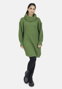 Vestido suéter verde de punto con cuello cisne, hombros caídos y textura acanalada. Llevado sobre leggings negros y botas negras gruesas.