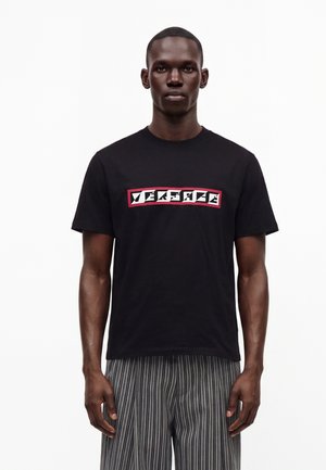 Homme portant un t-shirt noir Versace avec un logo blanc et rouge et un pantalon rayé gris, debout devant un fond blanc.