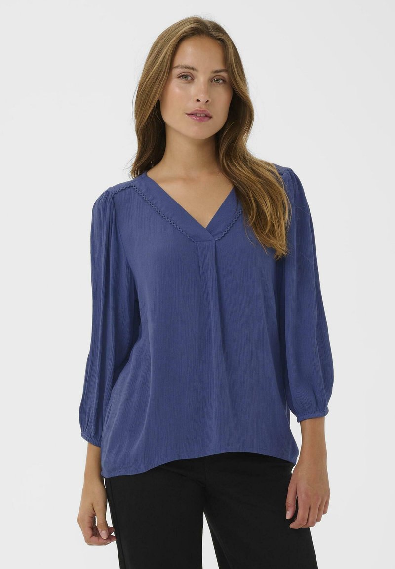 Blouse bleue à col en V avec des manches froncées, tissu texturé et couture décorative le long de l'encolure. Portée avec un pantalon noir.