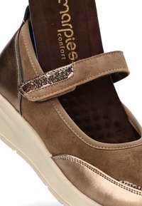 Zapato de cuero marrón con correa de velcro, superficie texturizada y plantilla acolchada. Presenta un diseño elegante y suela de goma de color claro.