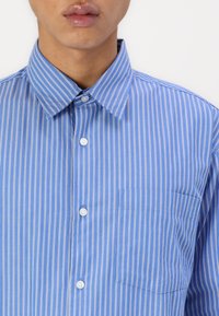 Blue striped button-up shirt with a collar, featuring white vertical stripes, a patch pocket, and white buttons, made from smooth fabric.