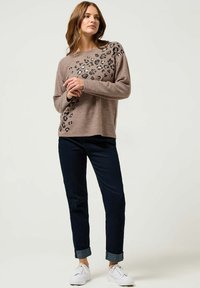 Brązowy sweter z czarnymi akcentami w panterkę, zestawiony z ciemnymi dżinsami i białymi sneakersami. Luźny krój i miękka tekstura.