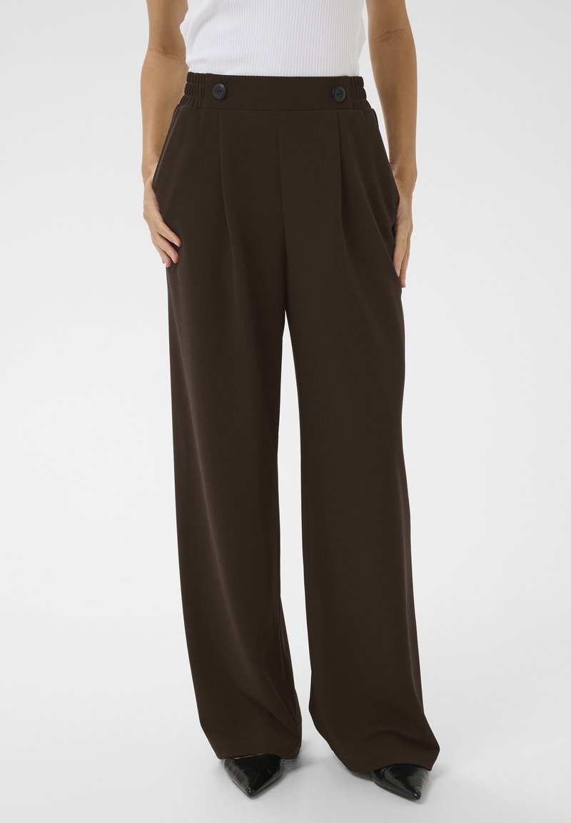 Femme portant un pantalon marron foncé taille haute à jambes larges avec des boutons à la taille et des chaussures noires pointues.