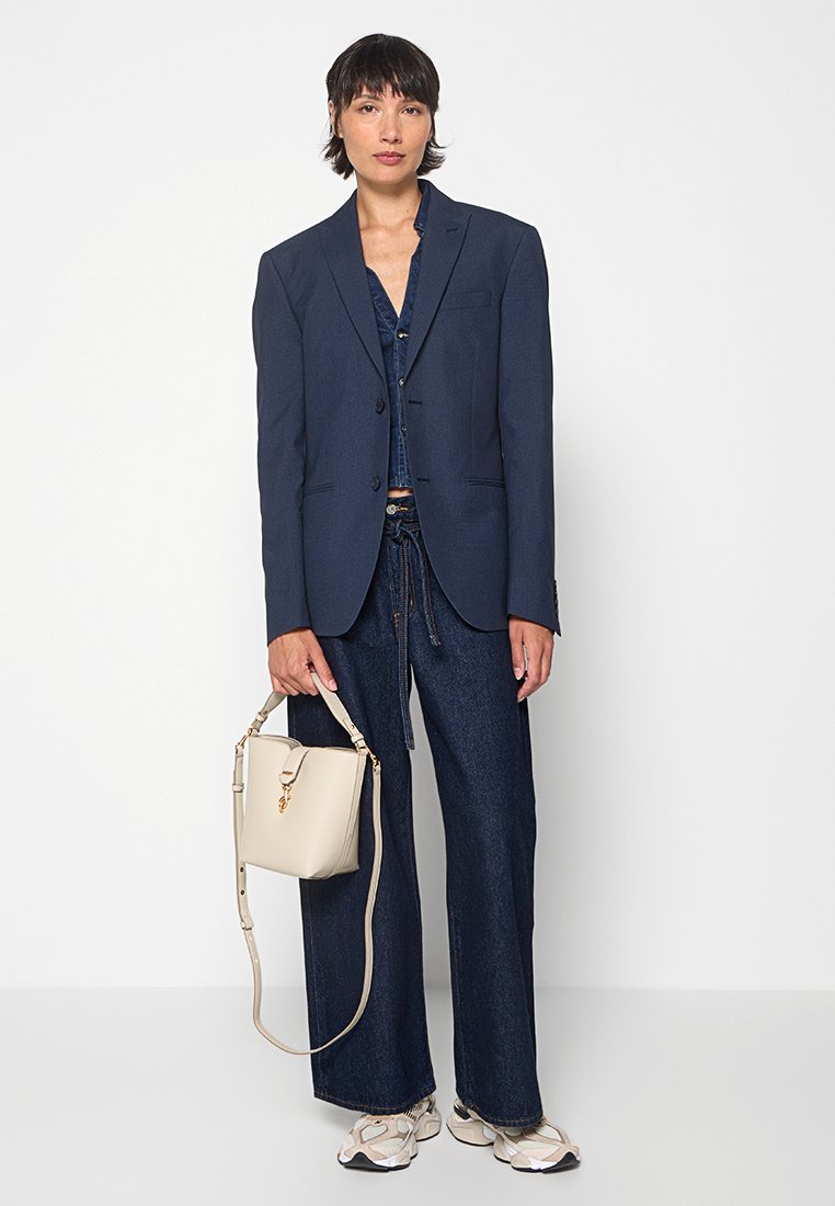 Marineblauer, taillierter Blazer mit einem einzelnen Knopf, kombiniert mit einem Jeanshemd und weit geschnittenen Jeans, ergänzt durch eine cremefarbene Handtasche und Sneakers.