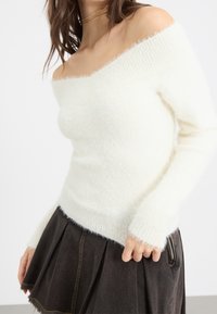 Maglione off-white, a spalla scoperta, realizzato in morbido e soffice materiale con collo a costine e orlo testurizzato, abbinato a una gonna plissettata scura.