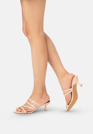 Nogi w beżowych lakierowanych sandałkach na niskim obcasie, z paskami i odkrytymi palcami, jedna stopa płasko na podłożu, druga uniesiona, na jednolitym jasnym tle.