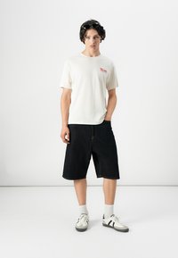 White cotton t-shirt with red text, paired with dark denim shorts. Model wears gray sneakers with black stripes and white socks.