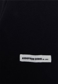 Tissu en maille noire avec une étiquette blanche portant le texte "REDEFINED REBEL RR - 2013." L'étiquette est positionnée le long du bord inférieur.