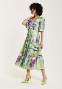 Maxi dress in vibrant green, blue, and purple abstract print. Flowy fabric, V-neckline, and short sleeves. Paired with bright green stilettos.