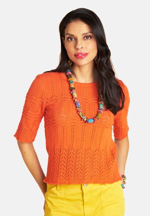 Woman wearing an orange patterned knit top, yellow pants, and colorful beaded necklace and bracelet, with long dark hair and neutral background.