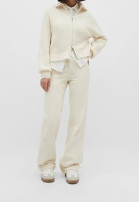 Un pull zippé côtelé beige clair associé à un pantalon large blanc cassé et des baskets blanches avec des accents beiges. Texture douce, coupe décontractée.